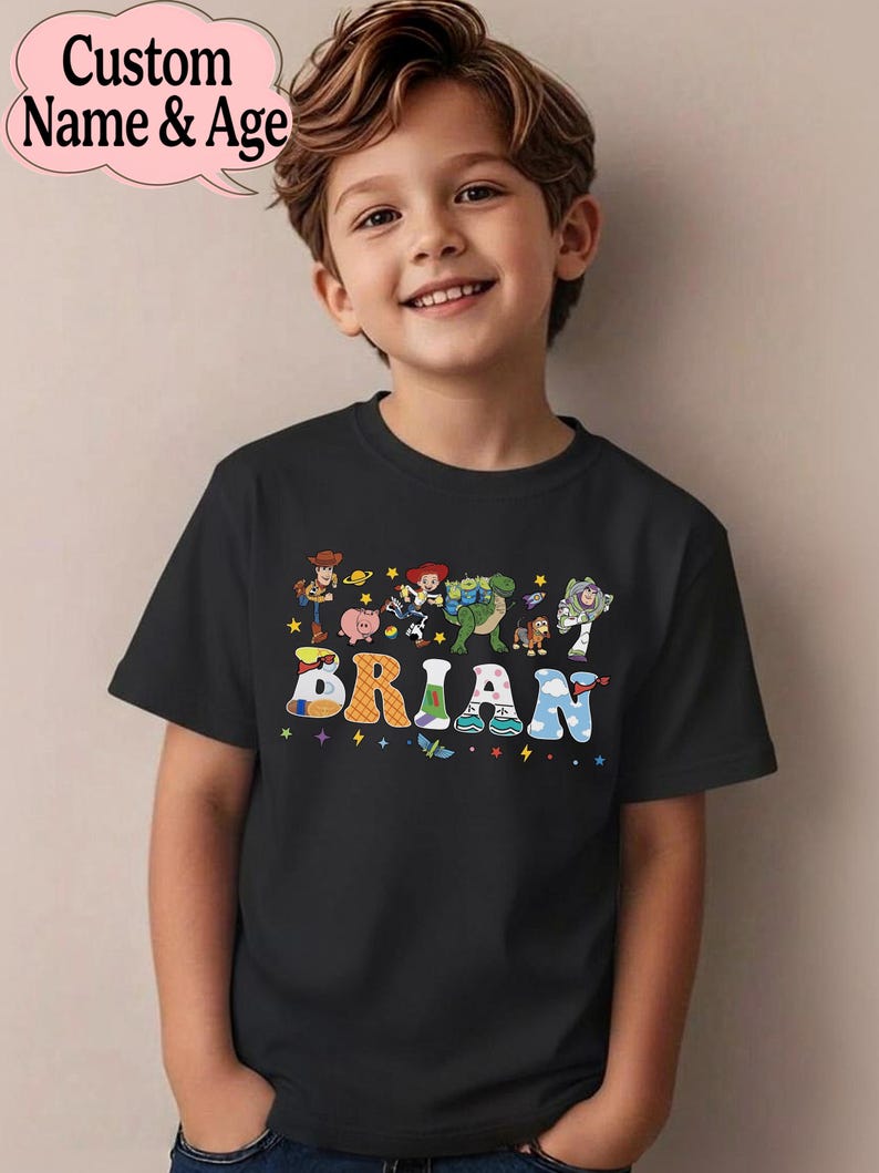 May include: A black t-shirt featuring the name "BRIAN" in colorful, playful letters. The shirt also displays cartoon characters. The words "Custom Name & Age" are in a pink bubble in the upper left corner.