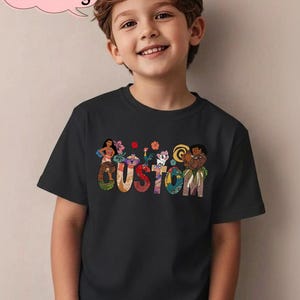 May include: Black t-shirt featuring the word "CUSTOM" in colorful, patterned letters. Above the word are cartoon characters and flowers. The shirt is worn by a smiling child. A pink speech bubble with the text "Custom Name & Age" is in the upper left corner.