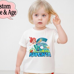 May include: White t-shirt featuring a colorful graphic of a mermaid, the number 6, and the name "Armenta" in blue. The shirt is designed for a child and is likely a birthday or special occasion garment.