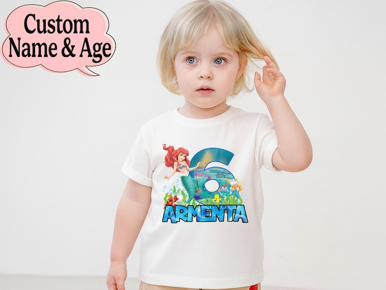 May include: White t-shirt featuring a colorful graphic of a mermaid, the number 6, and the name "Armenta" in blue. The shirt is designed for a child and is likely a birthday or special occasion garment.