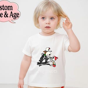 May include: White t-shirt featuring a cartoon graphic of Jack Skellington and other characters from The Nightmare Before Christmas. The word "nightmares" is printed below the graphic. A pink speech bubble with the text "Custom Name & Age" is in the top left corner.