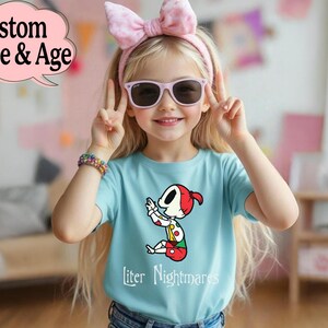 May include: A young girl wearing a light blue t-shirt with a cartoon character design and the text "liter Nightmares". She is wearing a pink bow headband, sunglasses, and a colorful bracelet. The image also includes the text "Custom Name & Age".