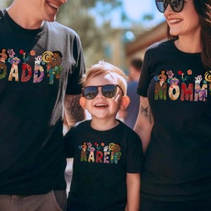 May include: Black t-shirts with colorful text and graphics. The shirts say "DADDY", "MOMMY", and "KAREN" in a decorative font with floral and character designs. The family is smiling and wearing sunglasses.
