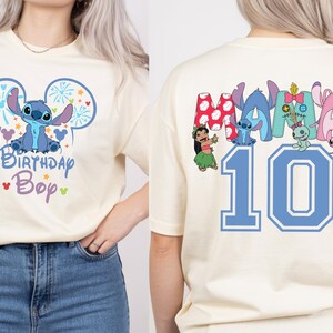 May include: Cream-colored t-shirt featuring a blue cartoon character with the text "Birthday Boy" on the front. The back of the shirt displays the number "10" in blue, along with various cartoon characters.