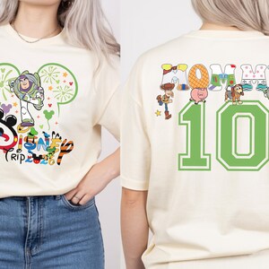 May include: Cream-coloured t-shirt with a Disney Trip 2023 graphic featuring Buzz Lightyear. The back of the shirt shows the word "Mommy" with Toy Story characters and the number "10" in green.