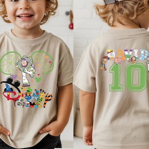 May include: A tan short-sleeved t-shirt with a colourful Disney Trip graphic featuring Buzz Lightyear and Mickey Mouse ears. The back of the shirt shows the name "DAVID" above the number "10" with Toy Story characters.