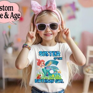 May include: A white t-shirt with a colorful graphic featuring the text "SISTER OF THE 6 BIRTHDAY GIRL" and a mermaid illustration. The girl is wearing sunglasses and a pink bow headband. A colorful bracelet is on her wrist.