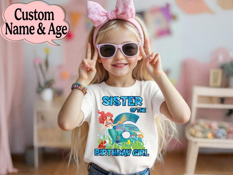 May include: A white t-shirt with a colorful graphic featuring the text "SISTER OF THE 6 BIRTHDAY GIRL" and a mermaid illustration. The girl is wearing sunglasses and a pink bow headband. A colorful bracelet is on her wrist.