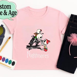 May include: A light pink t-shirt featuring a graphic of Jack Skellington and other characters from The Nightmare Before Christmas. The word "Nightmares" is printed below the graphic. The shirt is paired with dark blue jeans, a pink headband, and navy blue shoes.