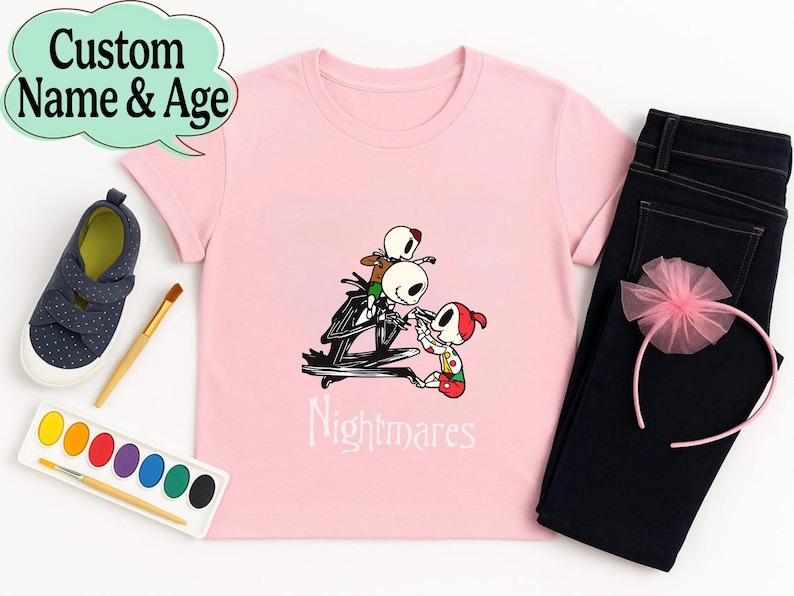 May include: A light pink t-shirt featuring a graphic of Jack Skellington and other characters from The Nightmare Before Christmas. The word "Nightmares" is printed below the graphic. The shirt is paired with dark blue jeans, a pink headband, and navy blue shoes.