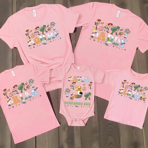 May include: Pink t-shirts and a onesie featuring a colorful graphic design with cartoon characters and the text "HAPPY BIRTHDAY". The shirts are short-sleeved and the onesie has short sleeves. The shirts and onesie are arranged on a wooden surface.