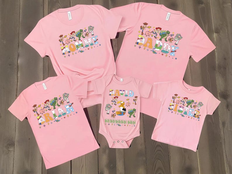 May include: Pink t-shirts and a onesie featuring a colorful graphic design with cartoon characters and the text "HAPPY BIRTHDAY". The shirts are short-sleeved and the onesie has short sleeves. The shirts and onesie are arranged on a wooden surface.