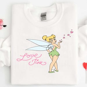 May include: White sweatshirt featuring a cartoon image of Tinkerbell blowing pink heart-shaped kisses. The words "Love Tink" are written in pink script. Pink heart-shaped sunglasses and heart-shaped lollipops are also visible.