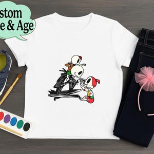 May include: White t-shirt featuring a cartoon illustration of Jack Skellington and other characters. The shirt is paired with dark blue jeans, a pink headband, and navy blue shoes. The text "Custom Name & Age" is in a speech bubble.