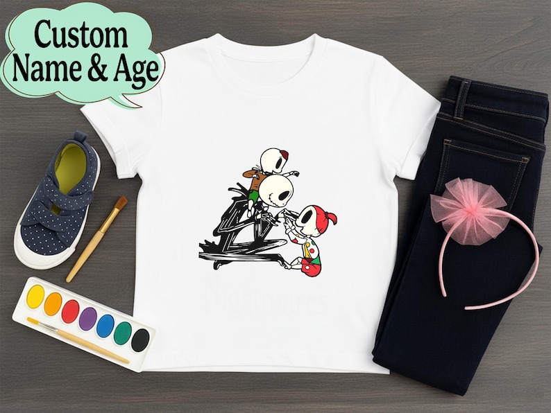 May include: White t-shirt featuring a cartoon illustration of Jack Skellington and other characters. The shirt is paired with dark blue jeans, a pink headband, and navy blue shoes. The text "Custom Name & Age" is in a speech bubble.