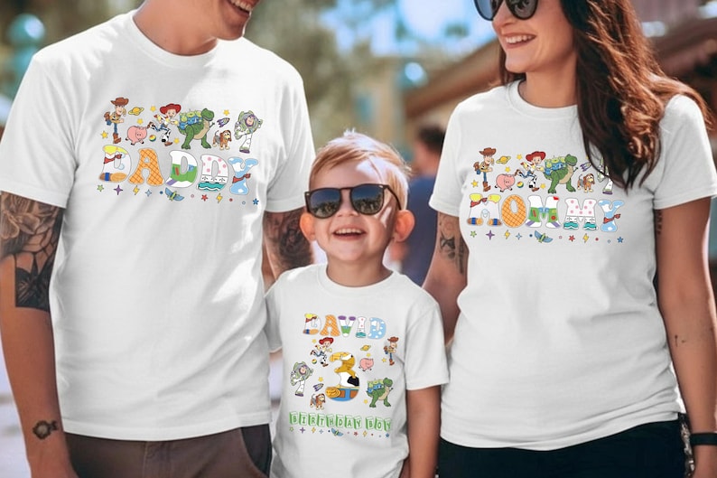 May include: White t-shirts with colorful Toy Story character designs. The shirts feature the words "DADDY", "MOMMY", and "DAVID" in playful fonts, with additional text "Happy Birthday Day". A family wears matching shirts.