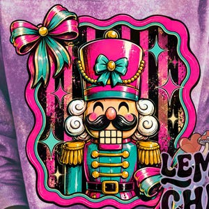 May include: A purple acid-wash sweatshirt featuring a graphic of a nutcracker with a mustache, a teal bow, and gold accents. The design includes a pink scalloped border, black and pink stripes, and the text Lemon Chill Design Co. with mushrooms.