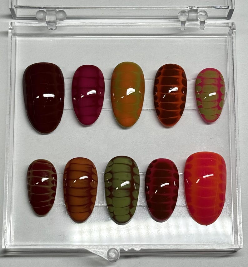 May include: A set of ten oval-shaped false nails in a clear plastic case. The nails feature a variety of colors, including burgundy, pink, orange, and green, with a textured, reptile-like pattern. The nails are designed for cosmetic use.