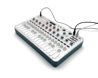Puede incluir: Dispositivo de producción de música electrónica con una superficie blanca y gris, con numerosas perillas, botones y una serie de almohadillas rectangulares. El dispositivo tiene un borde verde azulado oscuro y está conectado a cables negros. Una herramienta de producción musical moderna.