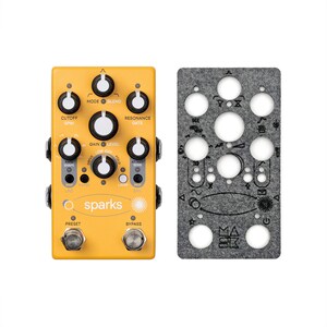 May include: A yellow Sparks guitar effects pedal with multiple knobs and switches, including "Cutoff," "Mode," and "Resonance." A gray felt cover with circular cutouts and symbols is next to the pedal. The pedal has the text "sparks" on it.