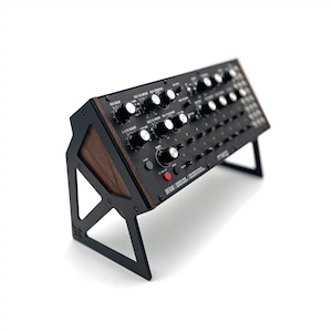 Op de afbeelding: Een zwarte Moog DFAM semi-modulaire analoge percussiesynthesizer gemonteerd op een zwarte metalen standaard met donkerhouten zijpanelen. Het bedieningspaneel is voorzien van talloze zilveren knoppen, zwarte knoppen, een rode knop en witte tekstlabels.