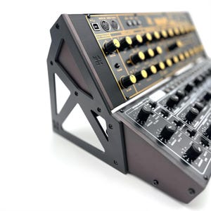 Puede incluir: Un sintetizador negro y dorado con numerosas perillas y botones. El sintetizador está montado en un soporte negro con un panel de madera. Los controles están etiquetados con texto blanco. El sintetizador está inclinado hacia el espectador.