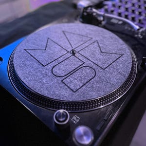 Puede incluir: Primer plano de un tocadiscos de DJ con un slipmat de fieltro gris con un diseño geométrico negro. El tocadiscos es negro con detalles plateados, incluyendo el brazo y los botones de control. El diseño es un logotipo estilizado.