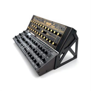 Puede incluir: Un sintetizador negro y dorado con numerosos botones y perillas. El texto "WASP DELUXE" es visible en el panel superior. El sintetizador está inclinado sobre un soporte negro, mostrando su diseño y características.