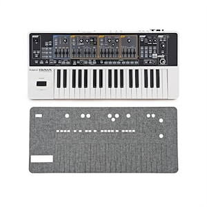 May include: A white Roland GAIA synthesizer with black and white keys and a control panel with various knobs and sliders. Below it is a gray felt cover with cutouts for the keys and controls.