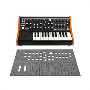 May include: A black synthesizer with white keys and numerous knobs and switches. The synthesizer has a wooden frame and the text "SUBSEQUENT 25" is visible. A gray felt cover with cutouts for the controls and keys is below.