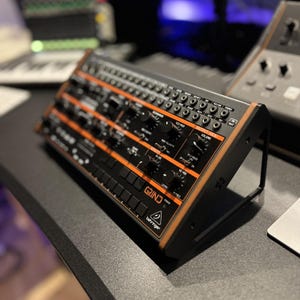 Könnte beinhalten: Ein schwarzer und orangefarbener Behringer Grind Synthesizer mit zahlreichen Knöpfen und Anschlüssen. Der Synthesizer hat einen Holzrahmen und ist nach oben abgewinkelt. Das Wort "GRIND" ist auf der Vorderseite aufgedruckt.