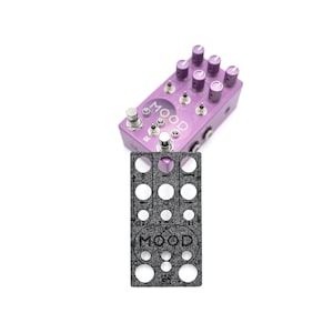 May include: A purple pedal with the word "MOOD" printed on it. The pedal has multiple knobs and a gray felt template with the word "MOOD" printed on it.
