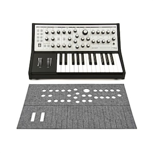 May include: A black and silver synthesizer with a keyboard and numerous knobs and switches. Below it is a gray felt mat with cutouts for the synthesizer's controls and keys. The synthesizer has the text "SUB PHATTY" printed on it.