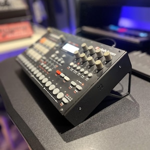 Op de afbeelding: Een zwarte Elektron Analog Rytm drumcomputer met een schuin metalen chassis, talloze lichtgrijze ronde knoppen, een centraal digitaal display en diverse draaiknoppen aan de rechterkant met gedrukte labels.