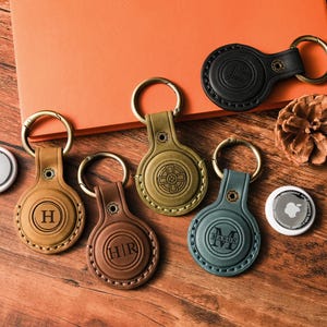 May include: A collection of leather keychains in various colors, including brown, olive green, and black, each with a gold-colored keyring. Some keychains feature embossed initials or designs. A white Apple AirTag is also present.
