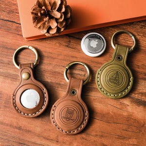 May include: Three brown leather AirTag holders with gold-colored key rings. One holder has an owl design, another has a sailboat, and the third has a blank circle. A white AirTag and a pinecone are also visible.
