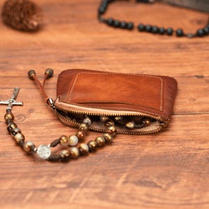 May include: A brown leather zippered pouch with a silver cross and brown beads inside. The pouch is open and the beads are spilling out.