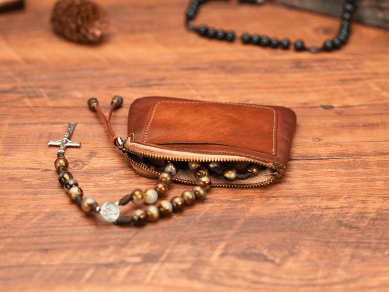 May include: A brown leather zippered pouch with a silver cross and brown beads inside. The pouch is open and the beads are spilling out.