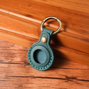 May include: A teal leather AirTag holder with a gold-colored key ring. The holder has a circular opening and a brass-colored button. The holder is on a wooden surface.