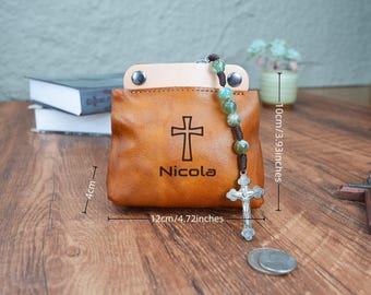 May include: A brown leather pouch with a cross and the name Nicola engraved on it. The pouch has a silver chain with a cross charm and green beads. The pouch measures 4.72 inches wide and 3.93 inches tall.