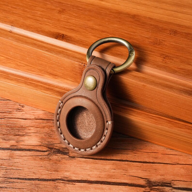 May include: A brown leather keychain holder with a brass-colored ring and button. The holder has a circular opening and is stitched with white thread. The background is a wooden surface.