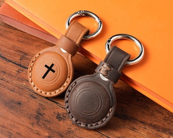 May include: Two leather keychains with silver rings. One is light brown with a black cross, the other is dark brown. Both have a circular design with stitched edges. The keychains are on a wooden surface.