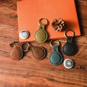 May include: A collection of leather keychains in various colors, including brown, green, and black, with gold-colored key rings. The keychains are designed to hold a small, round tracking device. A pinecone and an orange notebook are also in the image.