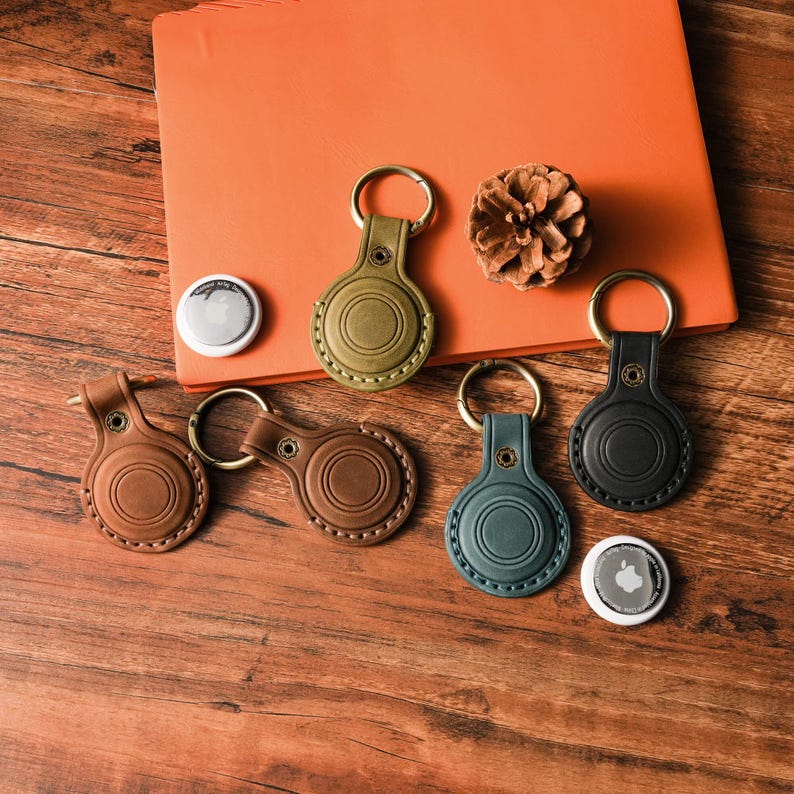 May include: A collection of leather keychains in various colors, including brown, green, and black, with gold-colored key rings. The keychains are designed to hold a small, round tracking device. A pinecone and an orange notebook are also in the image.