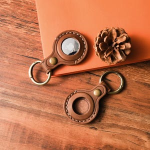 May include: Two brown leather AirTag holders with brass hardware and key rings are displayed on a wooden surface. One holder has a silver AirTag inside. A dried pine cone rests nearby on an orange surface.