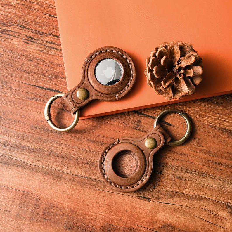 May include: Two brown leather AirTag holders with brass hardware and key rings are displayed on a wooden surface. One holder has a silver AirTag inside. A dried pine cone rests nearby on an orange surface.