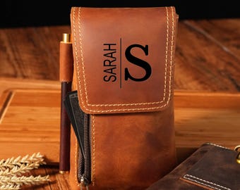 May include: A brown leather pen case with the name "SARAH" and the letter "S" printed on the front. The case has a zipper closure and a pen is visible in the side pocket. The case is sitting on a wooden surface.