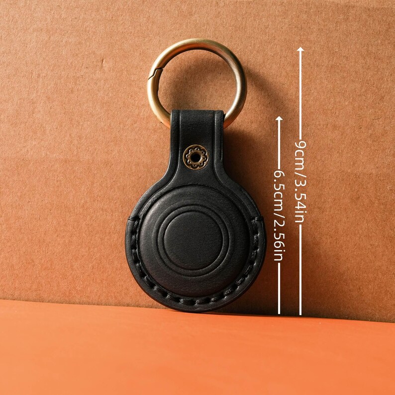 May include: Black leather keychain holder with a gold-colored ring. The holder is 3.54 inches long and 2.56 inches wide. The holder has a circular design with a stitched border and a decorative rivet.