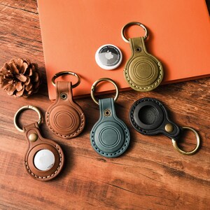 May include: A collection of leather AirTag keychains in various colors: brown, green, black, and olive green. Each keychain has a metal ring and a button closure. An Apple AirTag is also visible.