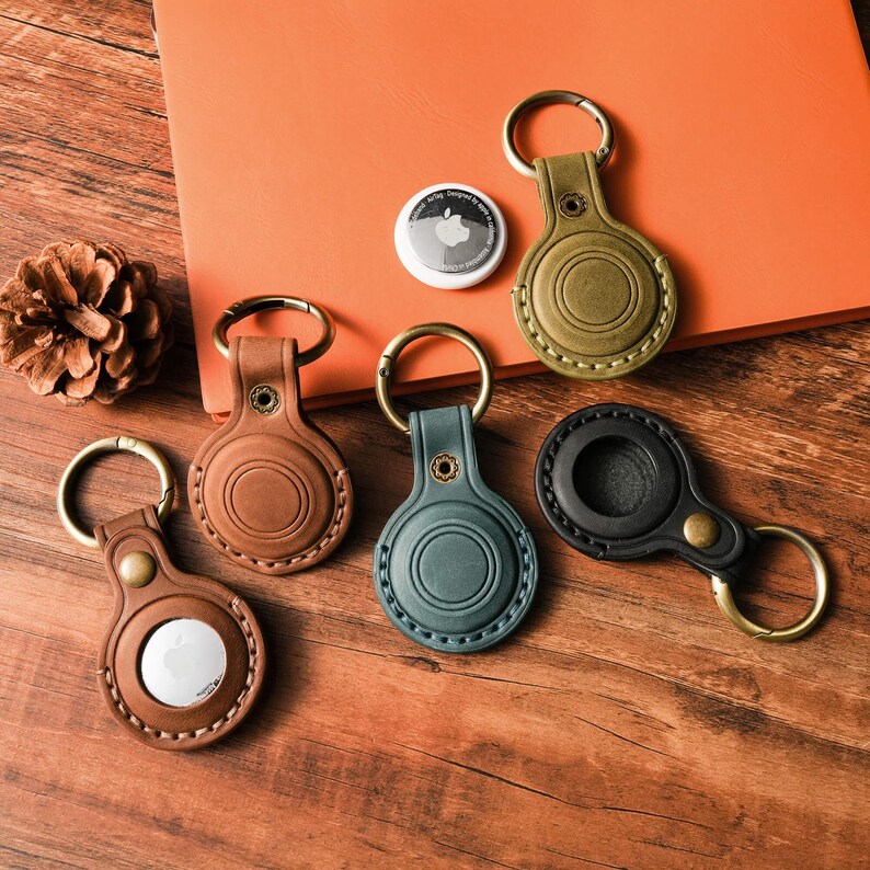 May include: A collection of leather AirTag keychains in various colors: brown, green, black, and olive green. Each keychain has a metal ring and a button closure. An Apple AirTag is also visible.